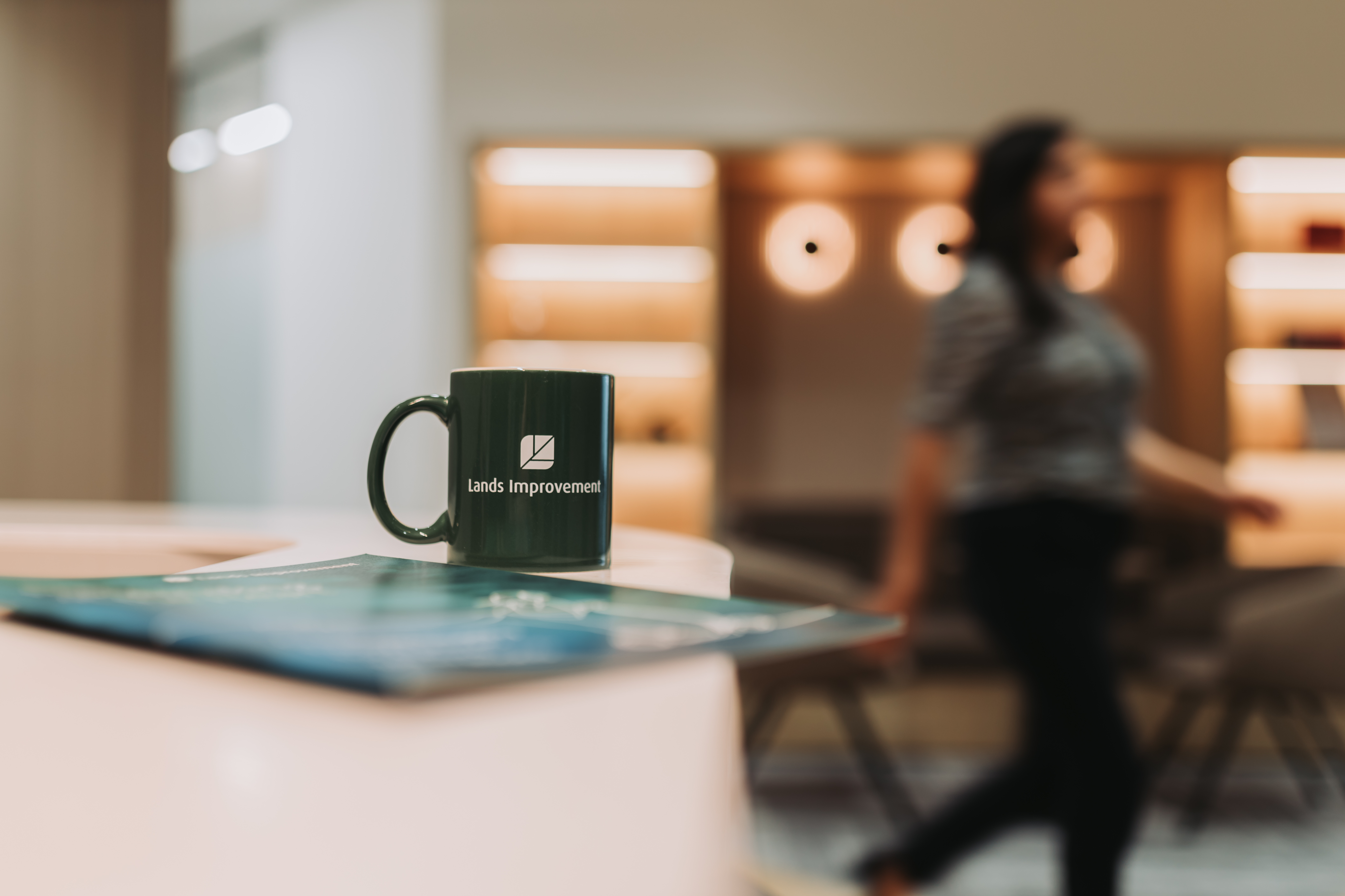 Black mug with white logo on blue surface, blurred office interior with orange lighting and person in background.