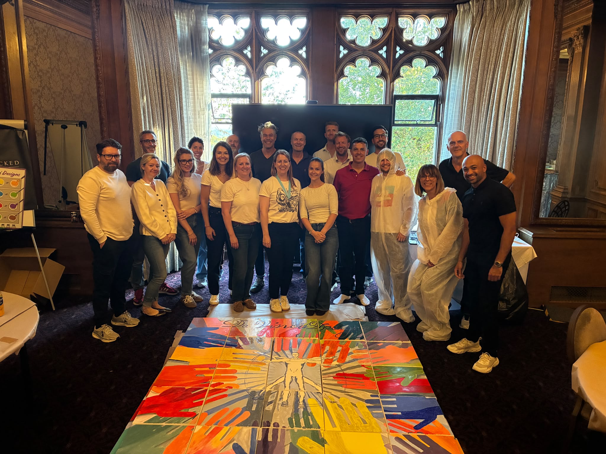 Large group of people standing behind a colourful painted canvas in a wood-panelled room with Gothic-style windows.