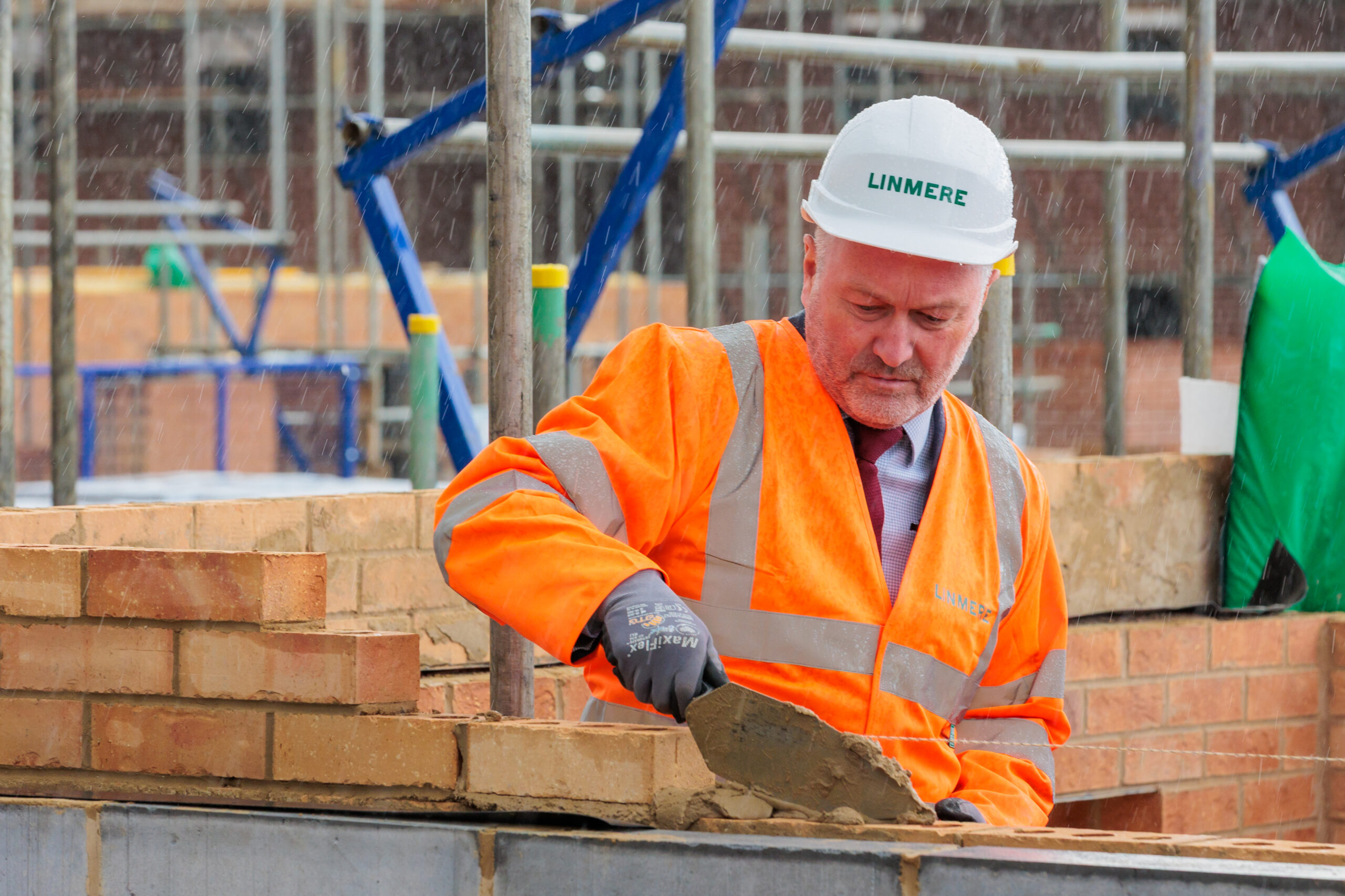 Construction worker in orange high-vis jacket and white hard hat laying bricks on building site with scaffolding in background.