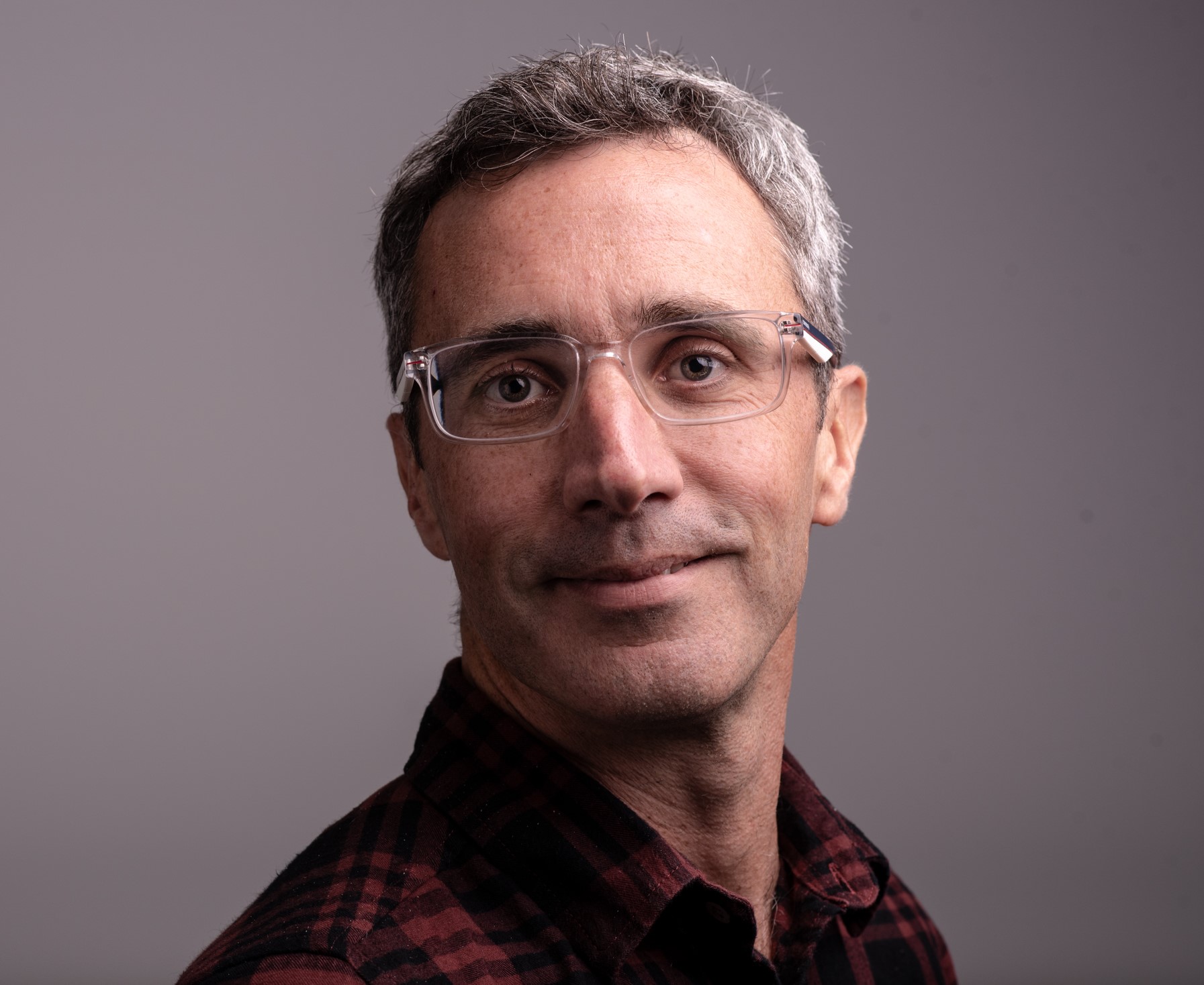 Middle-aged man with grey hair and clear-framed glasses wearing dark red checked shirt against grey background.