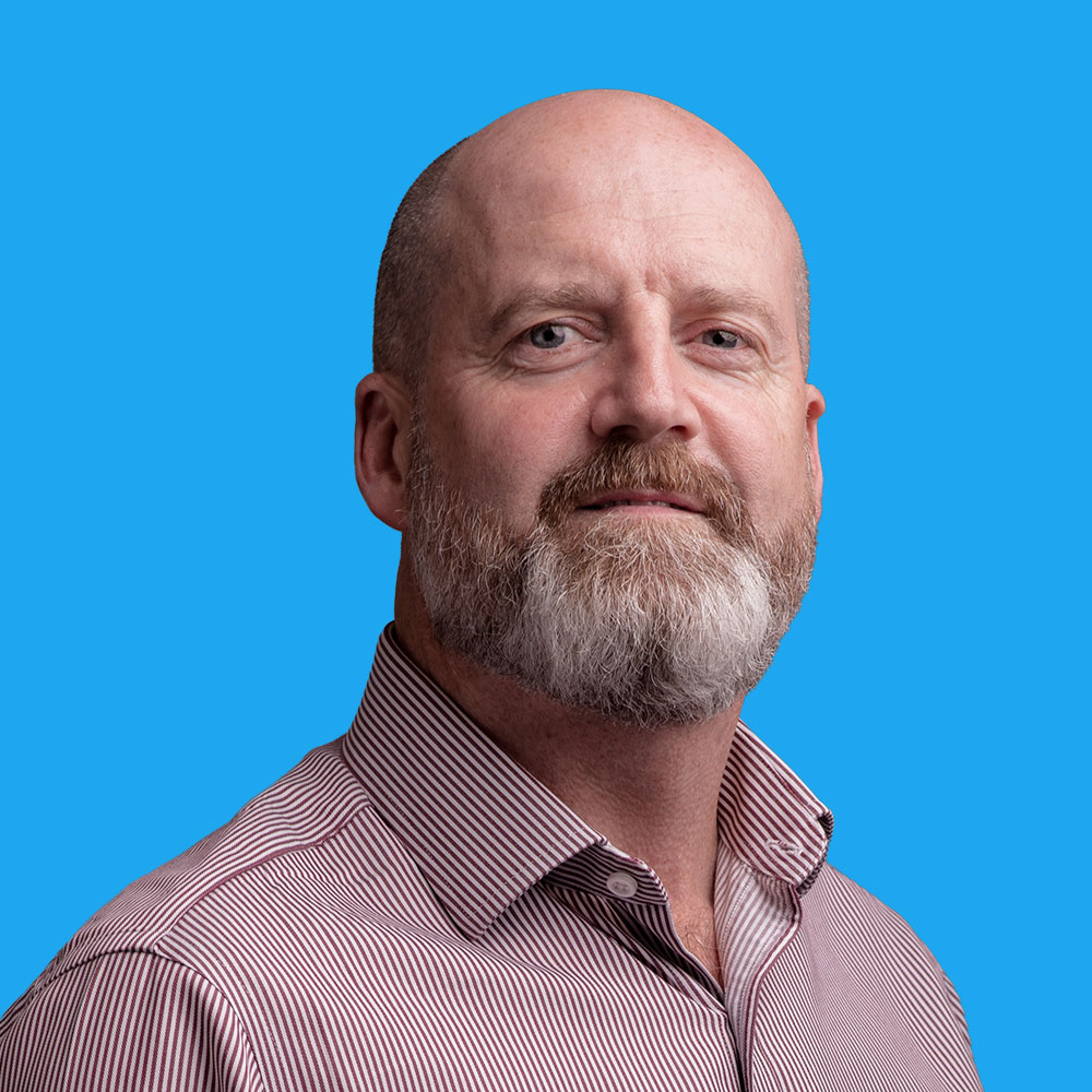 Bald man with grey beard wearing red and white striped shirt against bright blue background, smiling at camera.