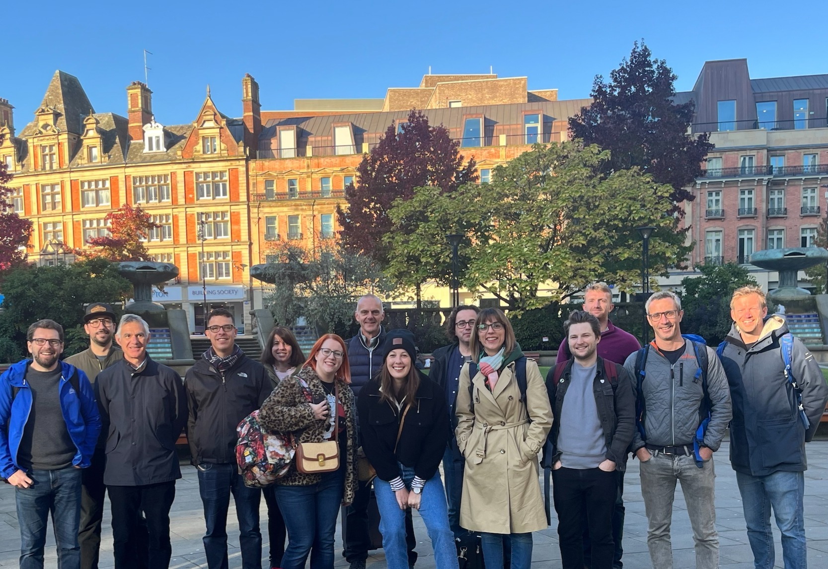 Group of people standing outdoors with colourful Victorian terraced buildings in background under blue sky with trees.