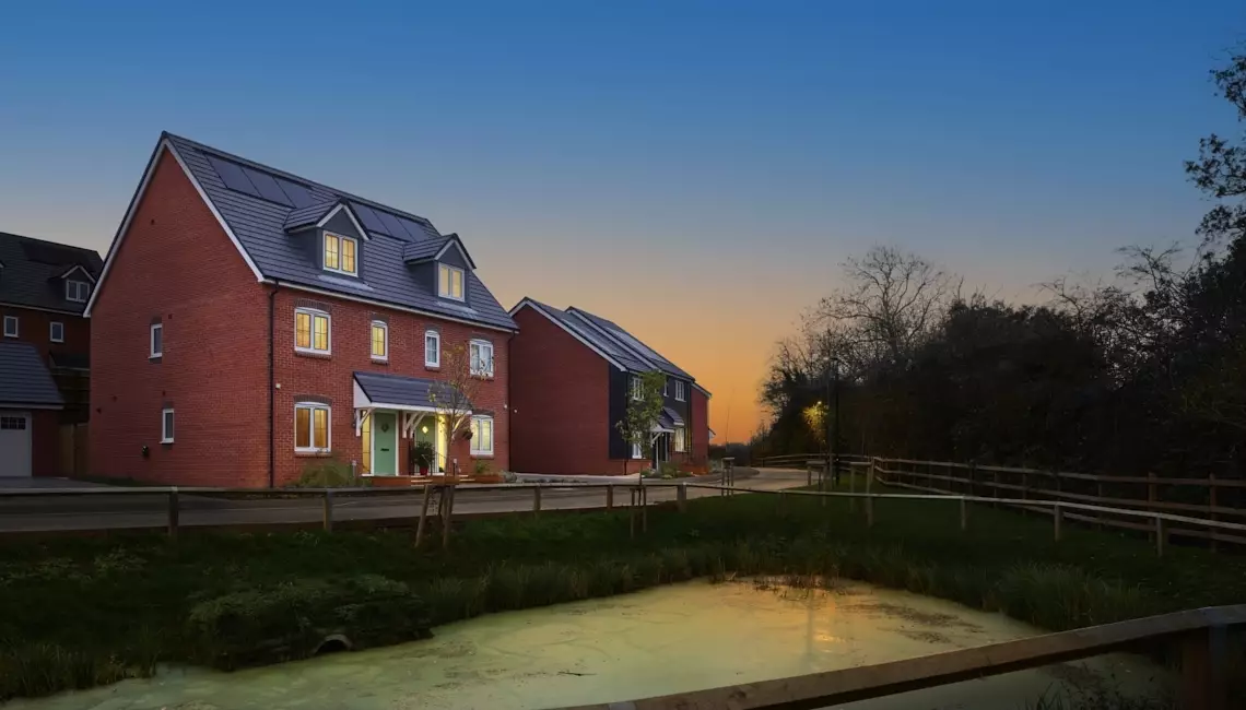 Two new-build red brick detached houses at dusk, with lit windows, solar panels, wooden fencing, and orange-blue twilight sky.