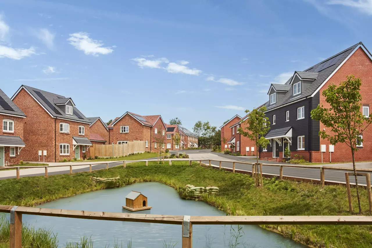 New-build red brick housing development with a small pond and duck house in the foreground, wooden fencing, and a tree-lined road under blue sky.
