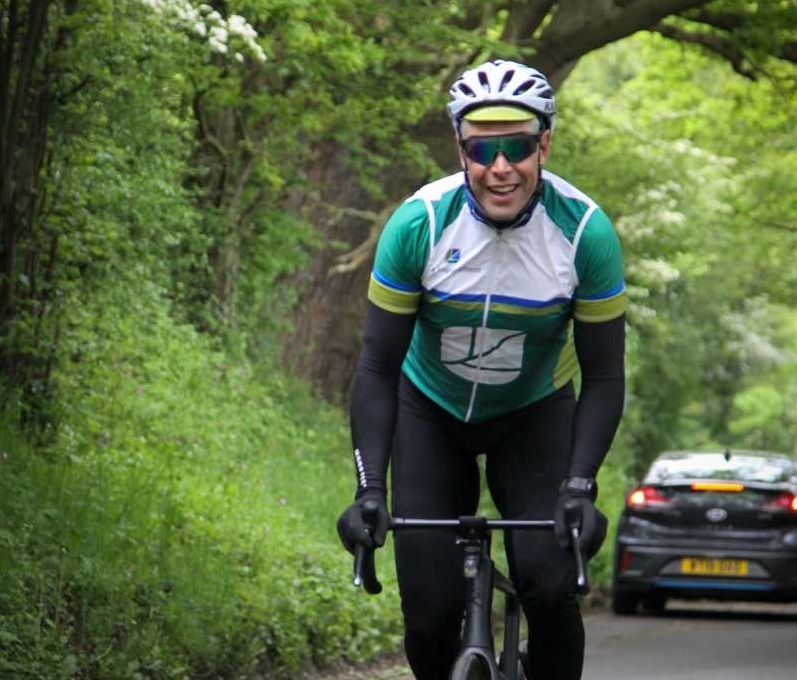 Cyclist in green and white jersey with helmet and sunglasses riding on tree-lined road with car behind in background.