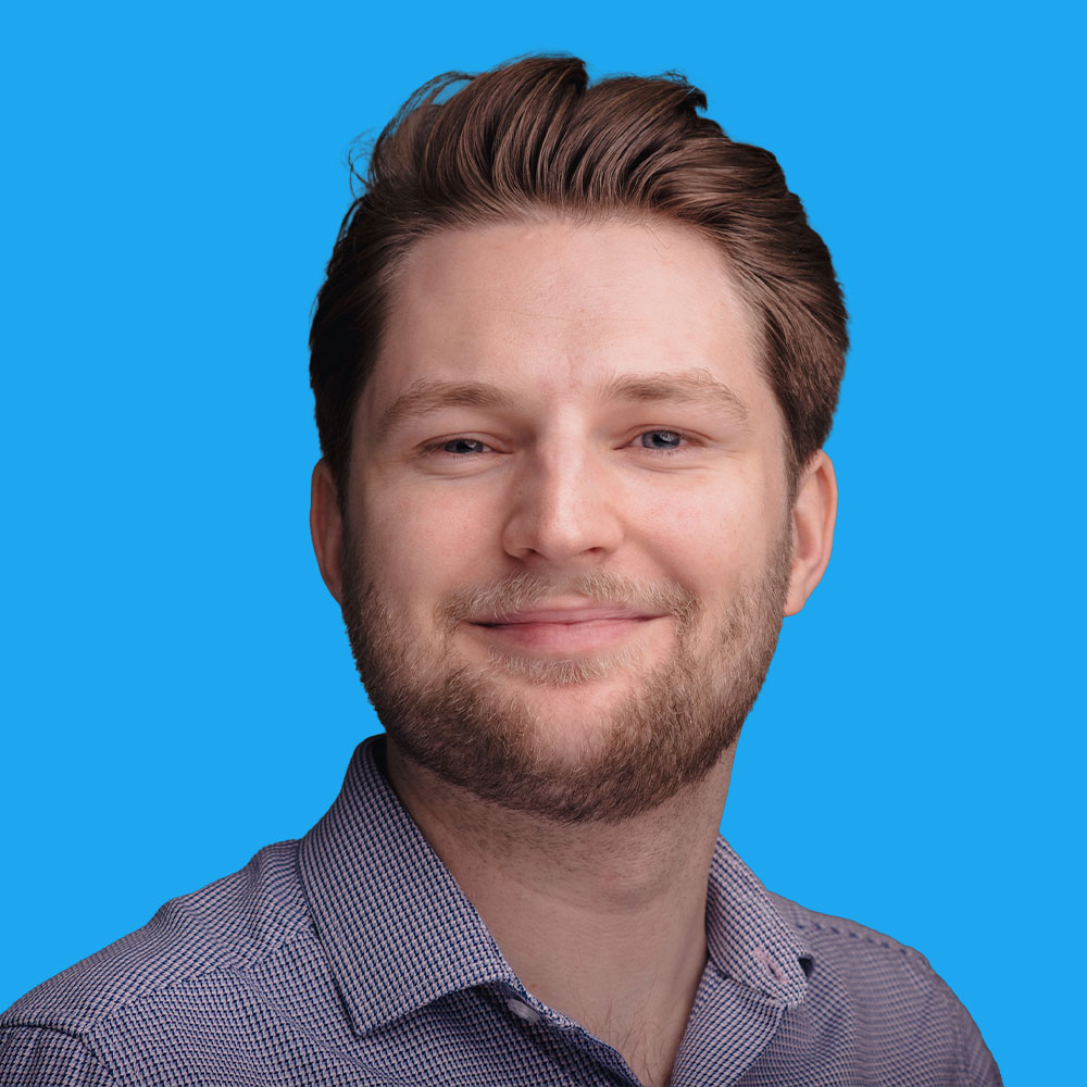 Man with brown hair and beard wearing striped shirt, smiling against bright blue background.