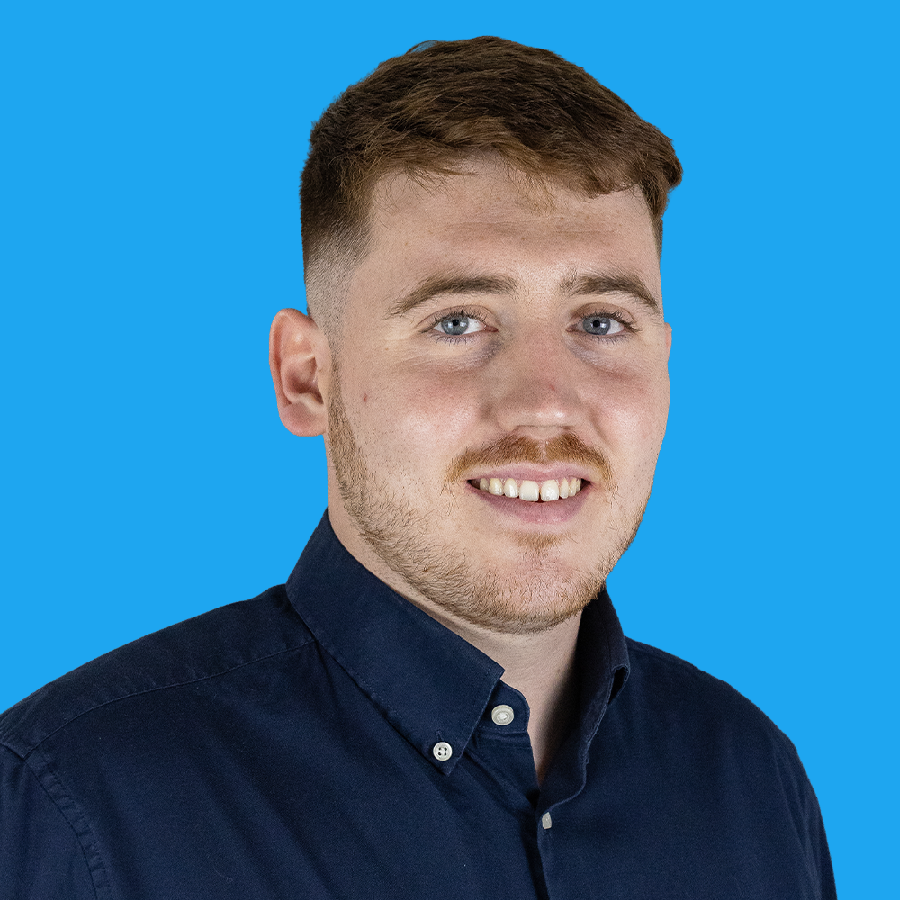 Young man with short brown hair and beard smiling, wearing dark blue shirt against bright blue background.