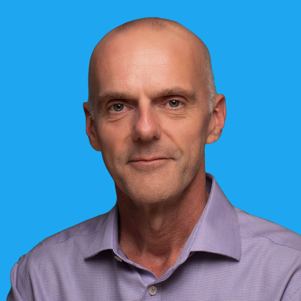 Bald middle-aged man in light purple striped shirt against bright blue background, facing camera with slight smile.