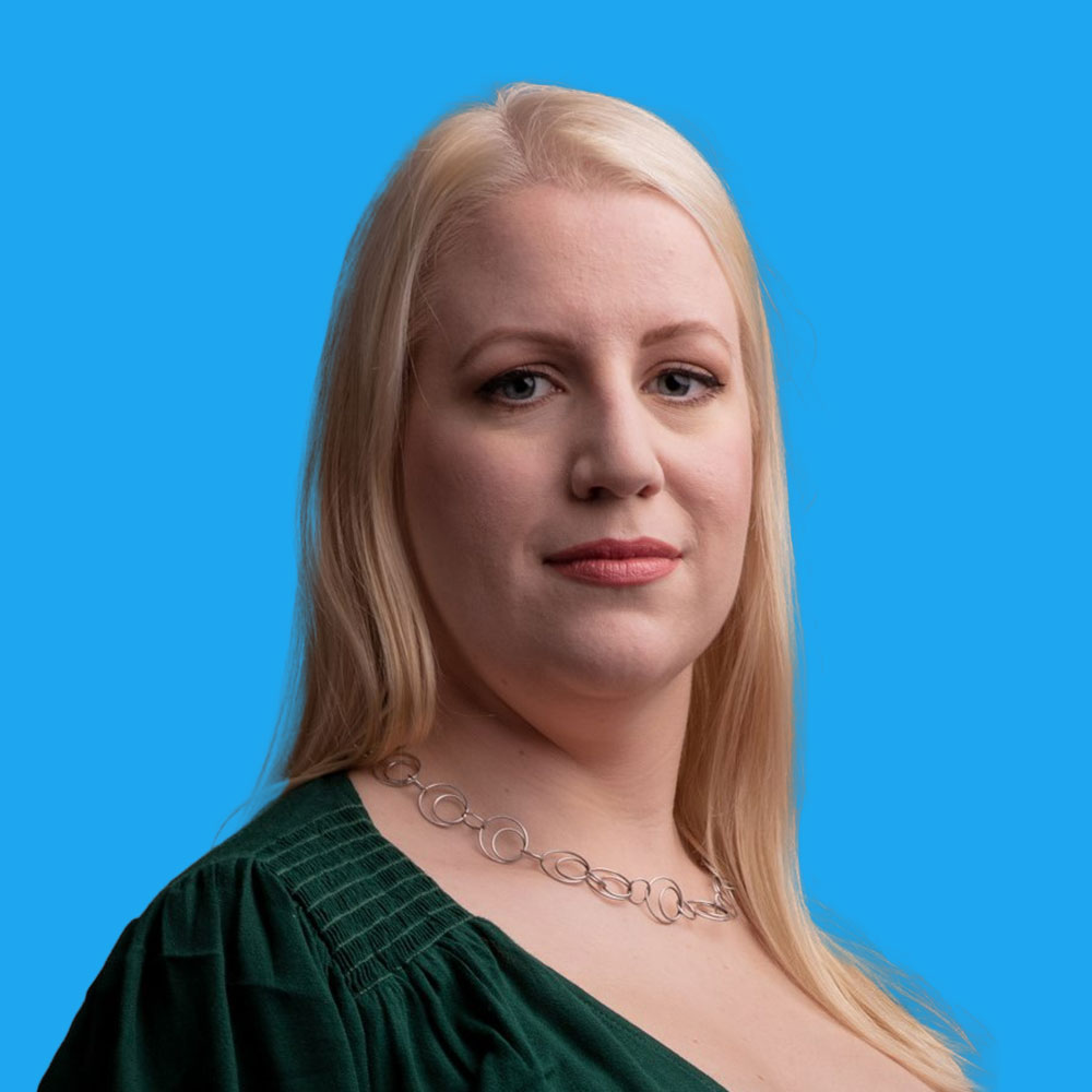 Blonde woman in dark green top with decorative necklace against bright blue background, facing camera with slight smile.