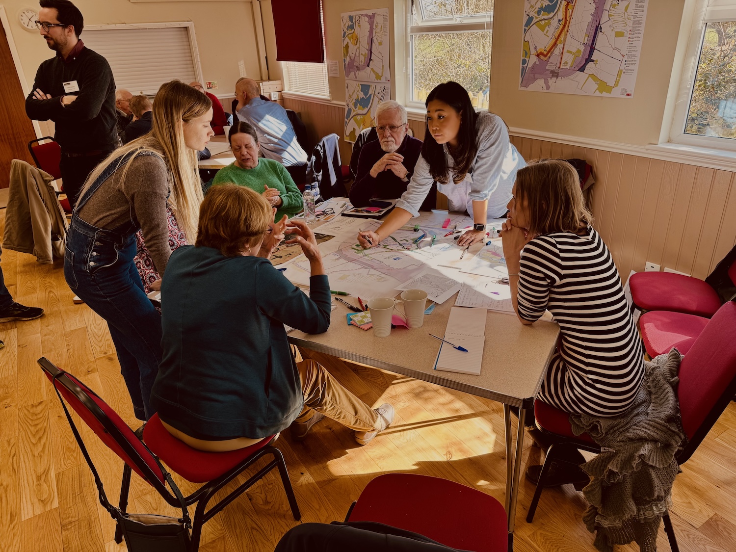 People gathered around white table with papers and materials in bright room with wooden floors and maps on walls.