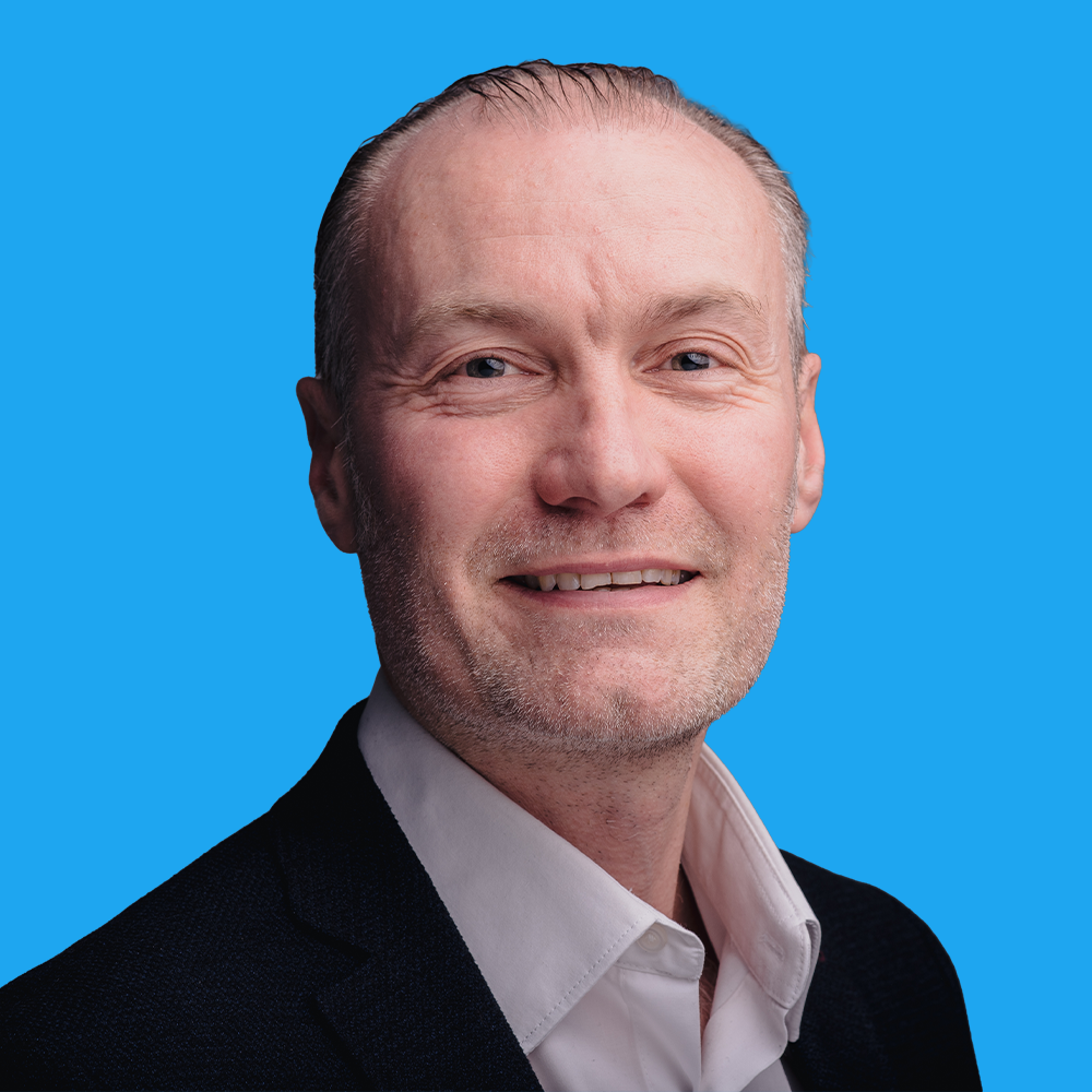 Middle-aged man with receding brown hair and stubble, wearing dark blazer over white shirt, smiling against bright blue background.