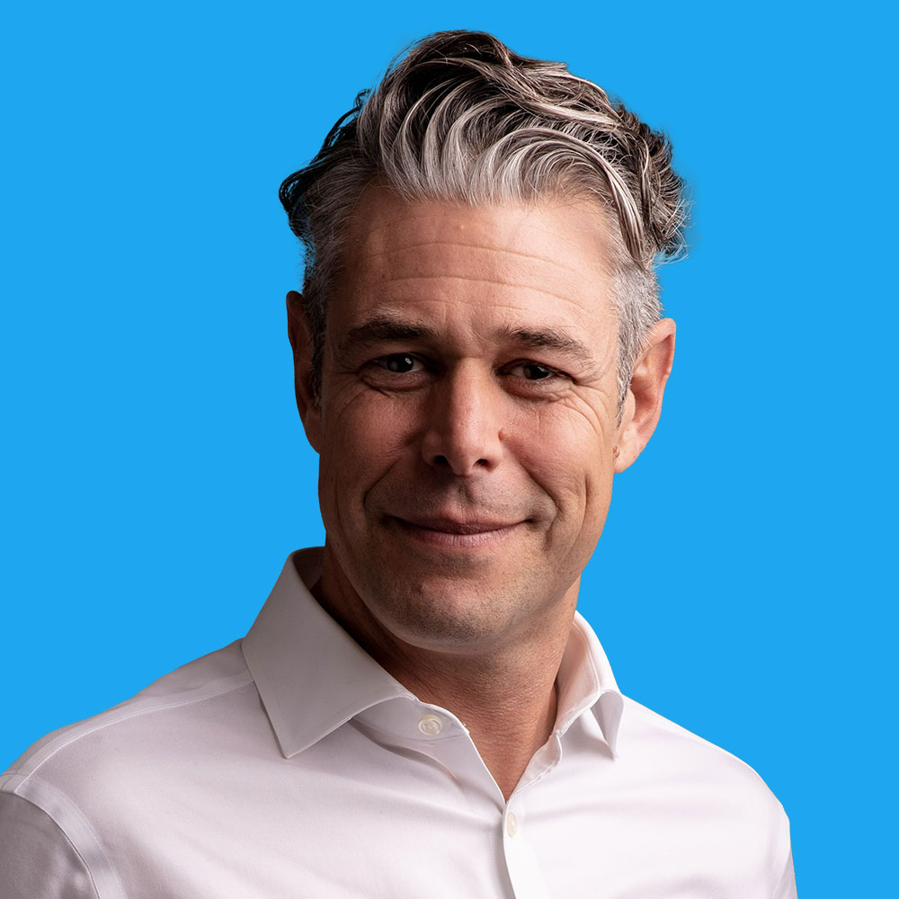Middle-aged man with grey-blonde hair wearing white collared shirt against bright blue background, smiling at camera.