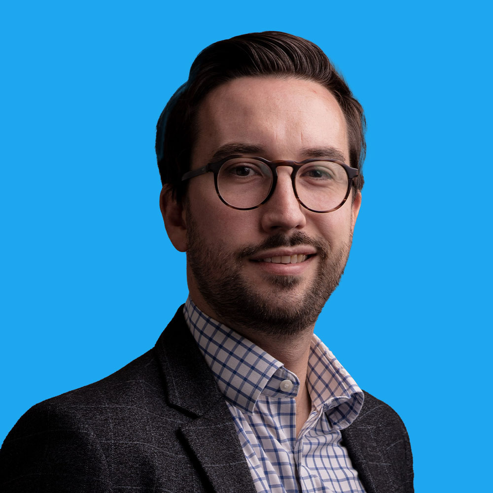Man with beard wearing glasses, dark jumper over checked shirt, smiling against bright blue background.
