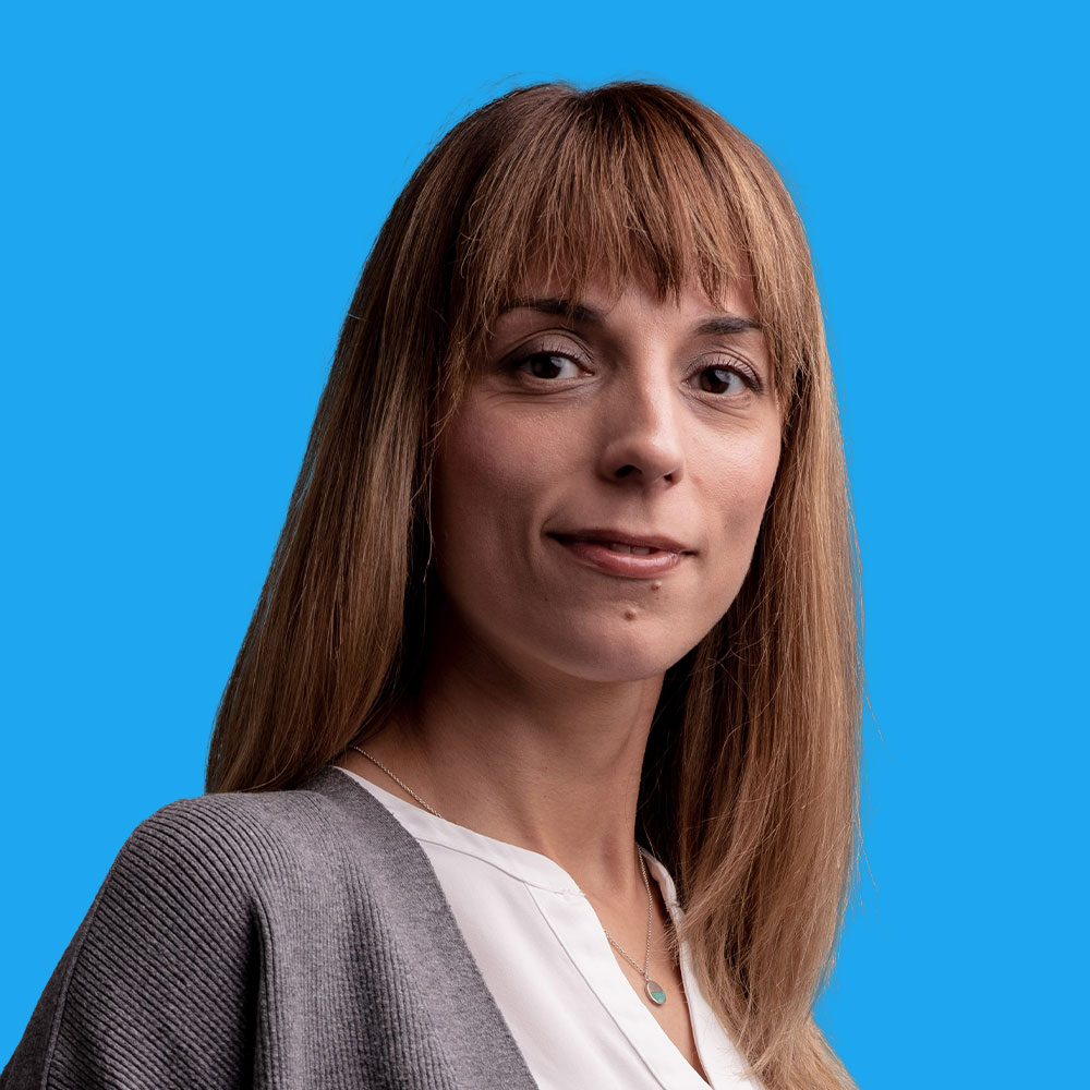 Woman with blonde fringe and long hair wearing grey blazer over white top, smiling against bright blue background.