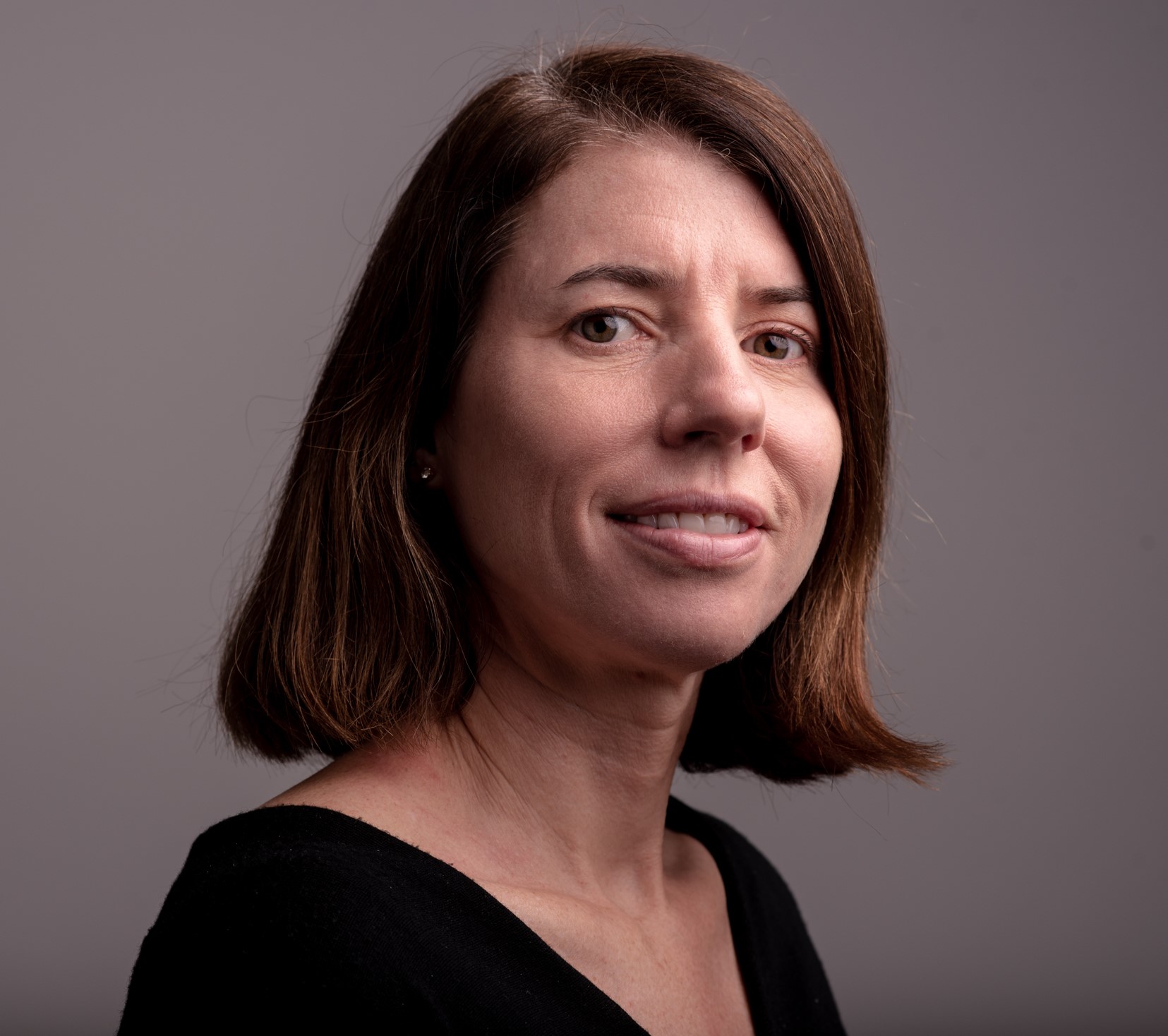 Woman with shoulder-length brown hair wearing black top, smiling at camera against grey background.