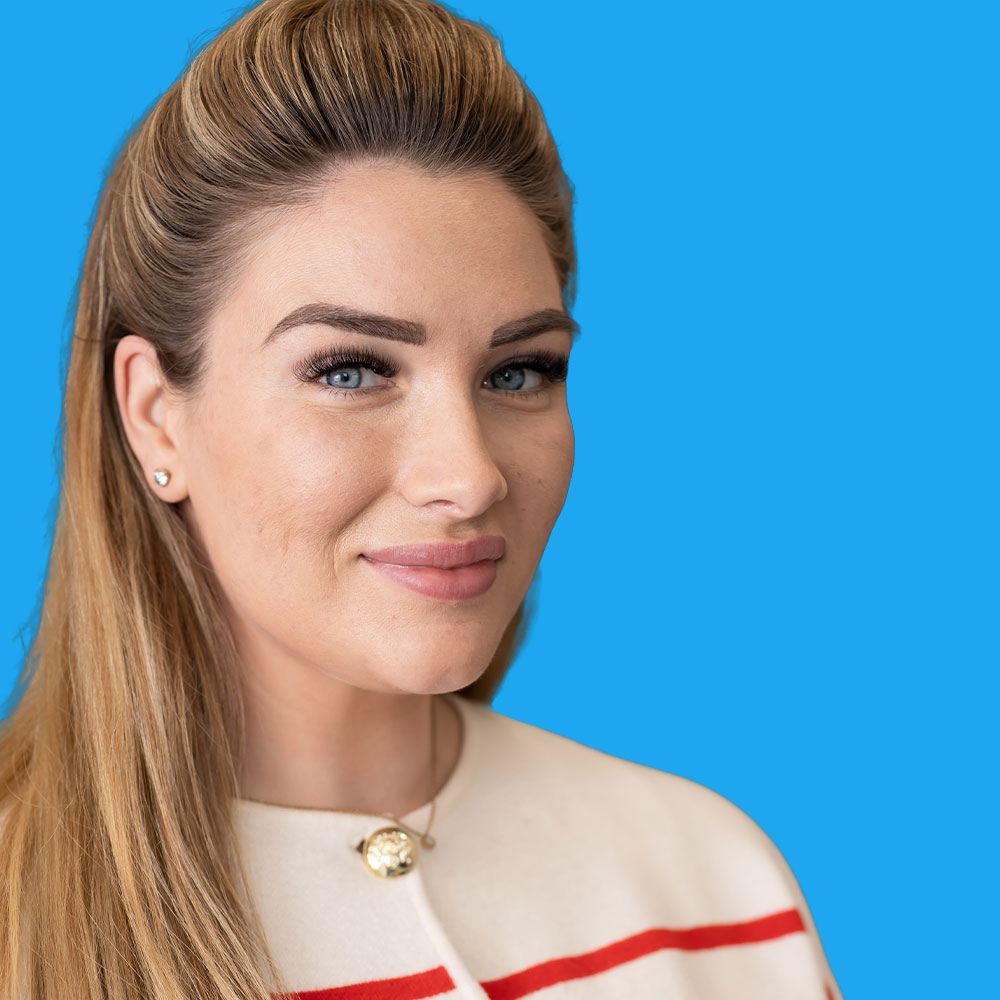 Woman with blonde hair pulled back wearing white top with red stripe, smiling against bright blue background.