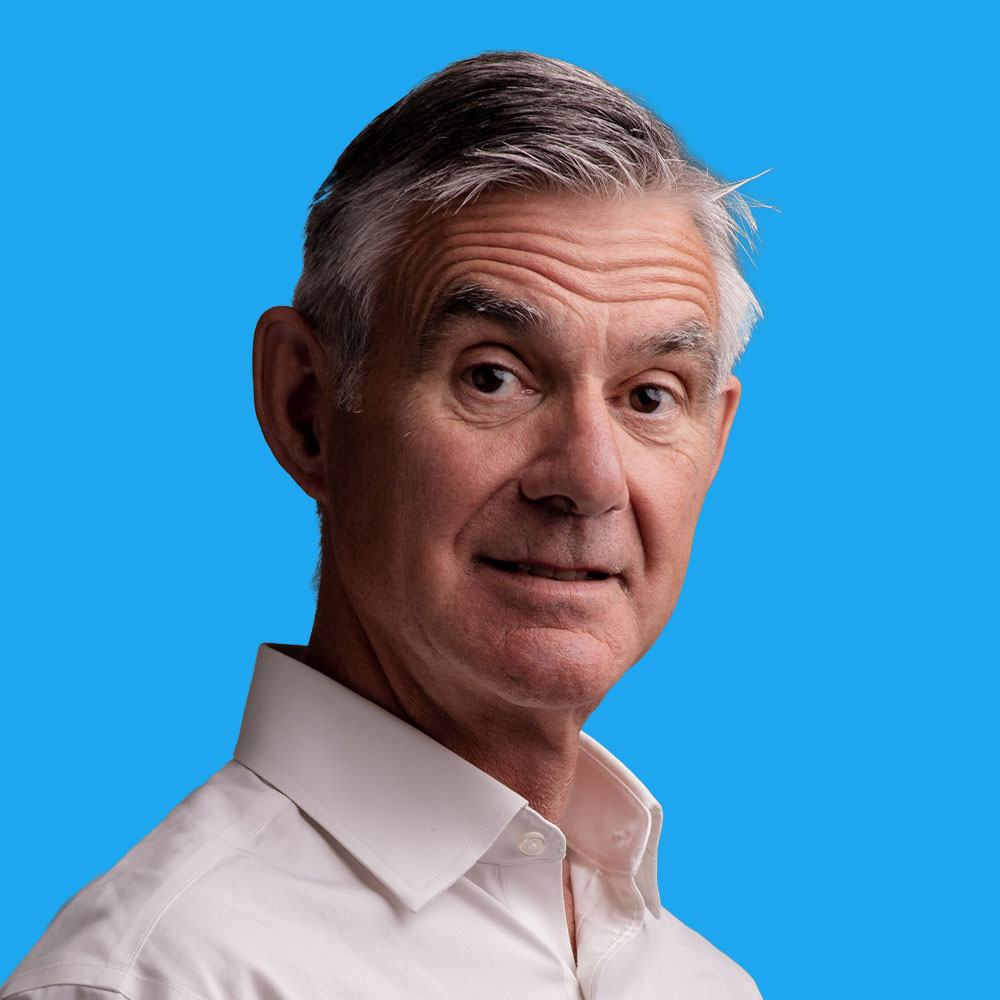 Middle-aged man with grey hair wearing white collared shirt against bright blue background, looking at camera with slight smile.