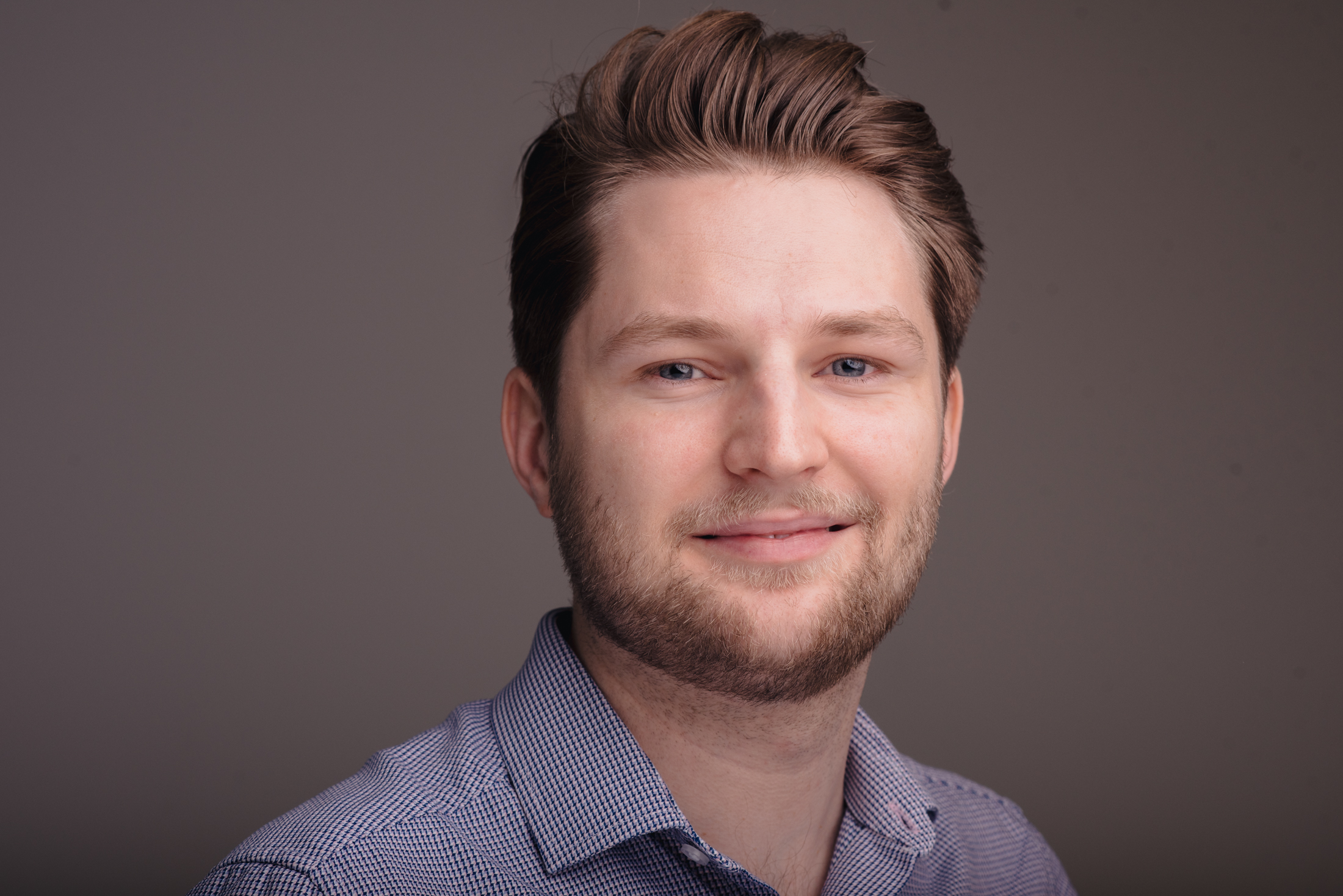 Man with brown hair and beard wearing blue checked shirt, smiling at camera against grey background.