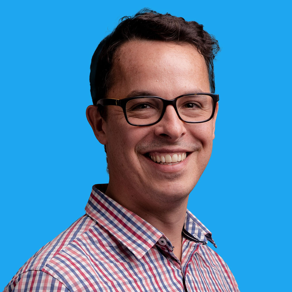 Smiling man with dark-rimmed glasses wearing red and blue chequered shirt against bright blue background.