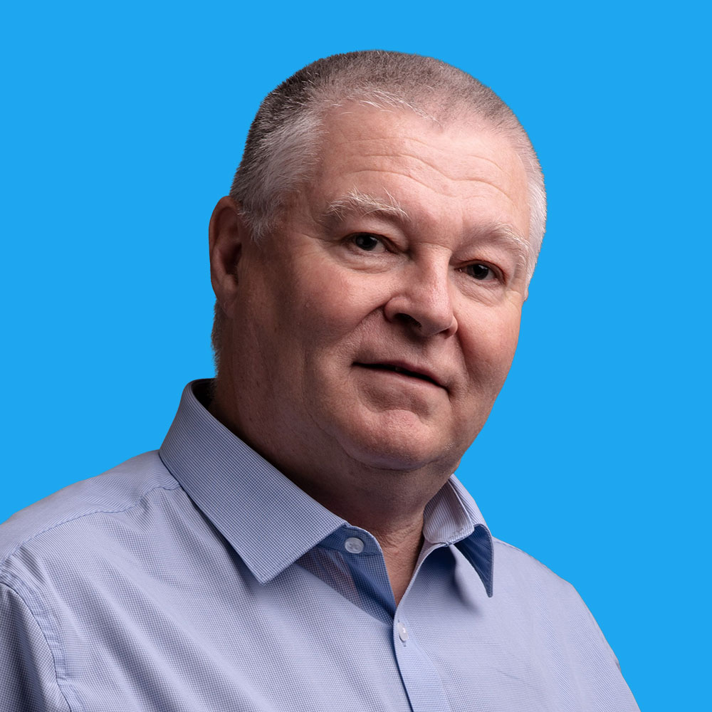 Middle-aged man with short grey hair wearing light blue button-up shirt against bright blue background, looking directly at camera.