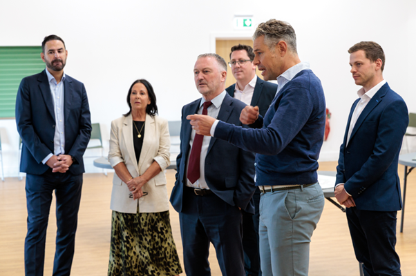 Group of seven people in business attire standing in modern office space with white walls and wooden flooring.