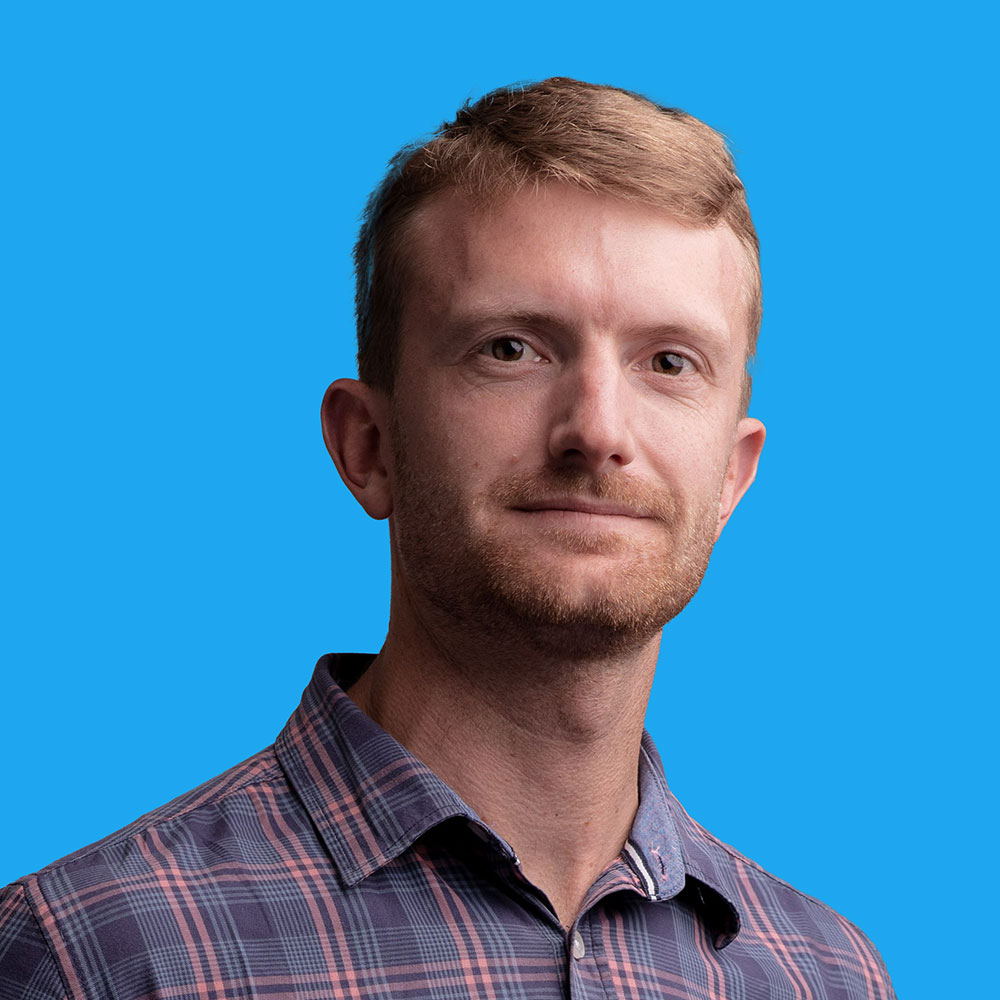 Man with short brown hair and beard wearing grey checked shirt against bright blue background.