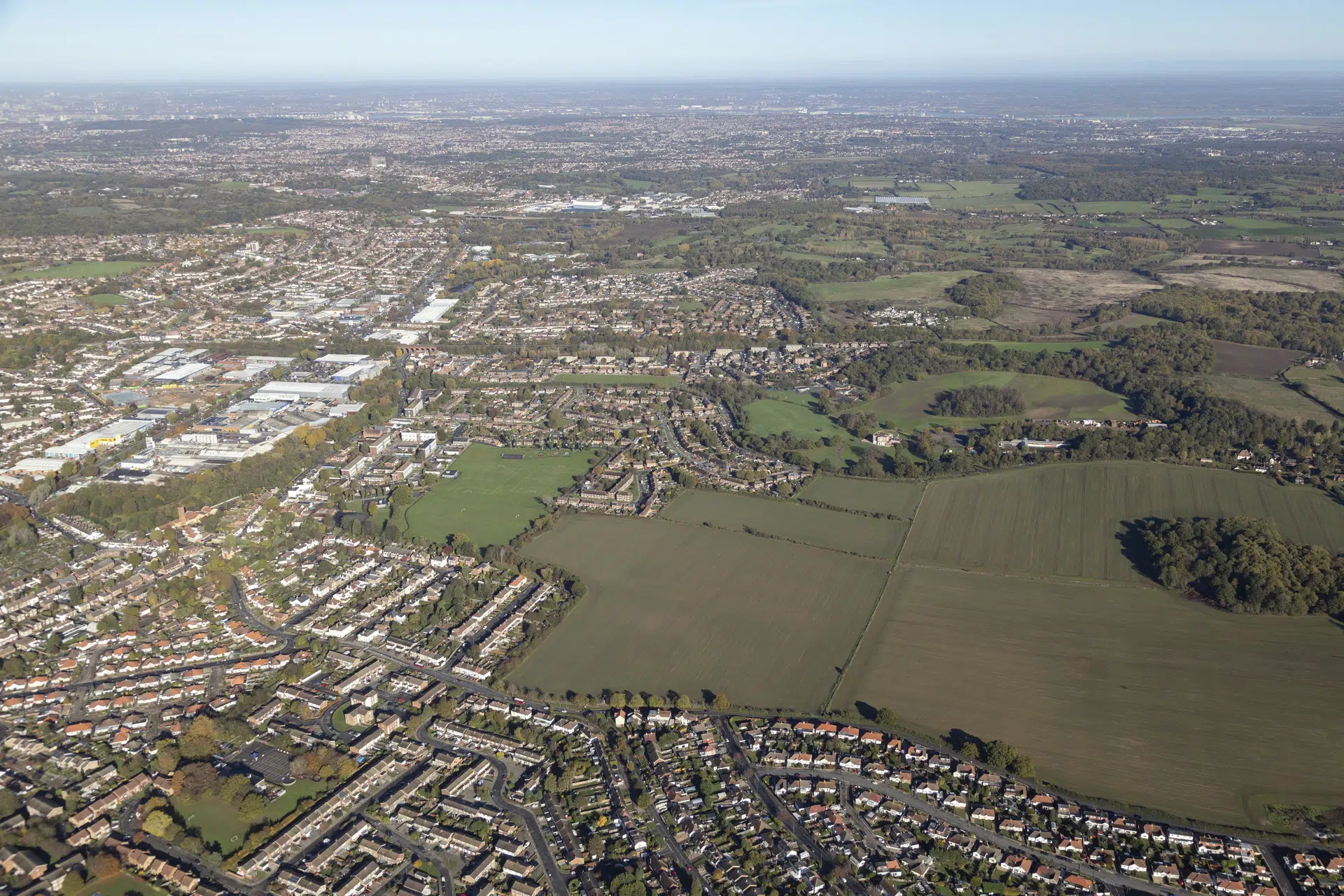Aerial view of suburban housing estates with geometric street patterns, green fields, and wooded areas under hazy sky.