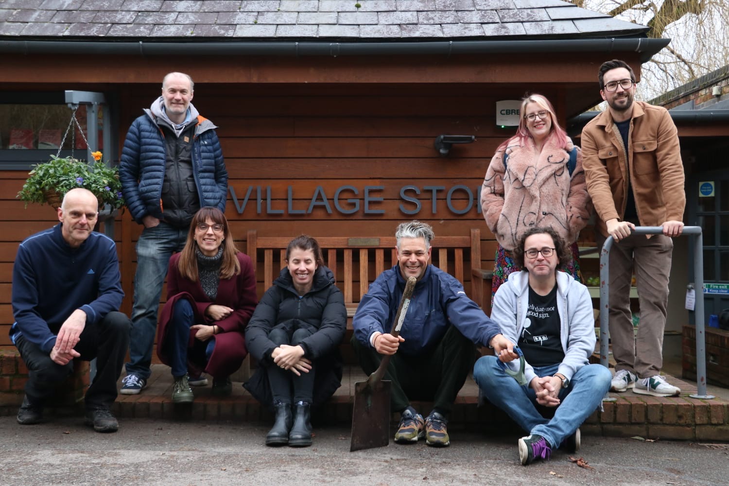 Eight people posing outside Village Store, mix of standing and sitting on low wall, wearing winter jackets and casual clothing.