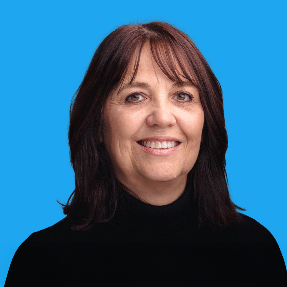 Woman with shoulder-length brown hair and fringe smiling at camera, wearing black top against bright blue background.