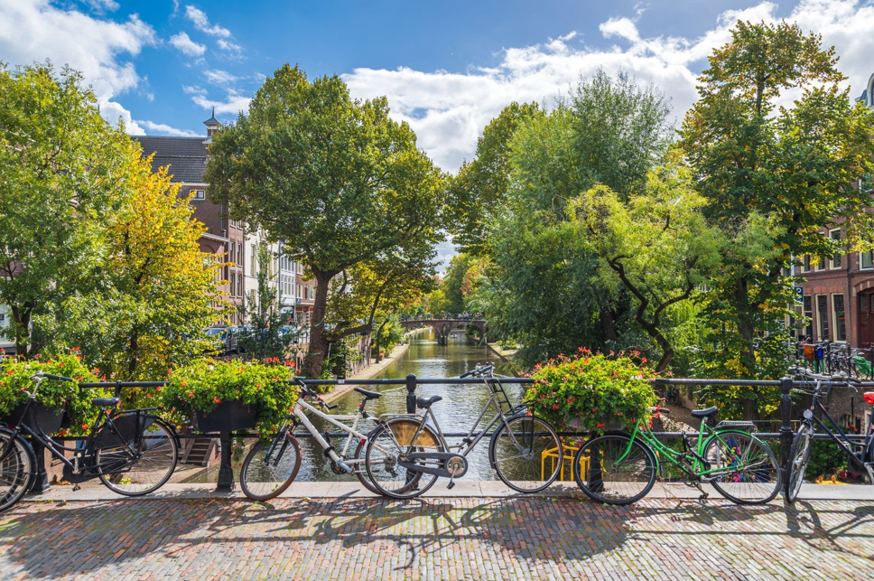 Bicycles parked on bridge railing overlooking canal with boats, surrounded by leafy trees and traditional brick buildings under blue sky.