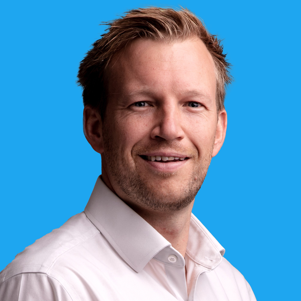 Man with brown hair and stubble wearing white collared shirt, smiling against bright blue background.