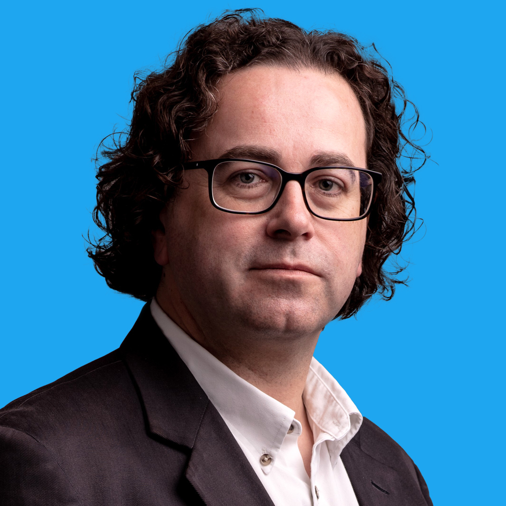 Man with curly brown hair and glasses wearing dark suit jacket and white shirt against bright blue background.