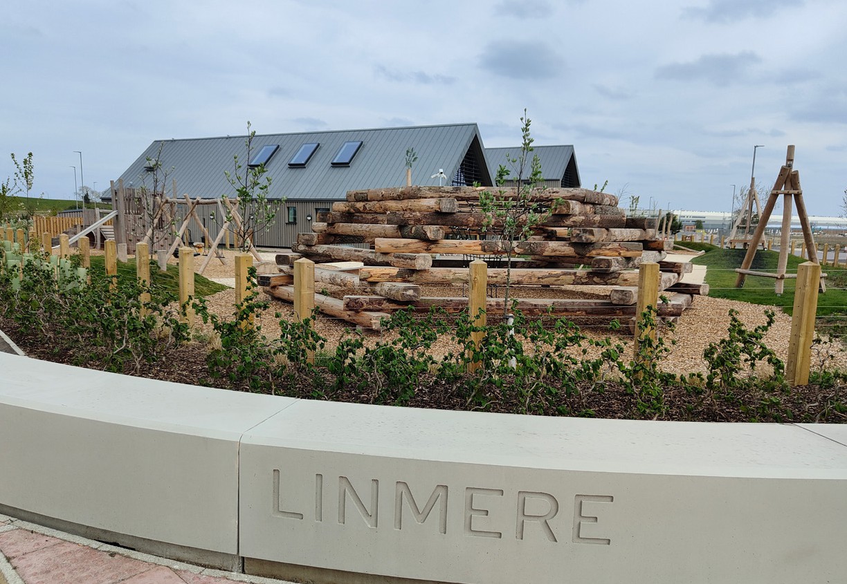 Large timber logs stacked behind wooden fence with "LINMERE" engraved on concrete wall. Modern house with grey roof in background.