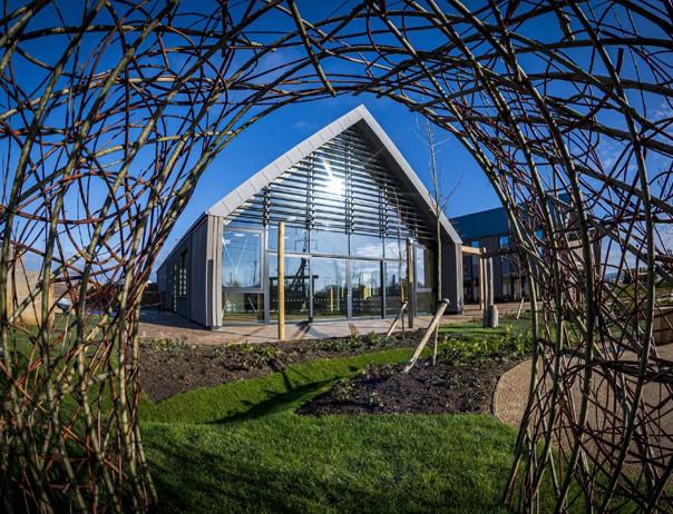 Modern glass-fronted building with triangular roof viewed through circular woven branch structure, green lawn, blue sky.