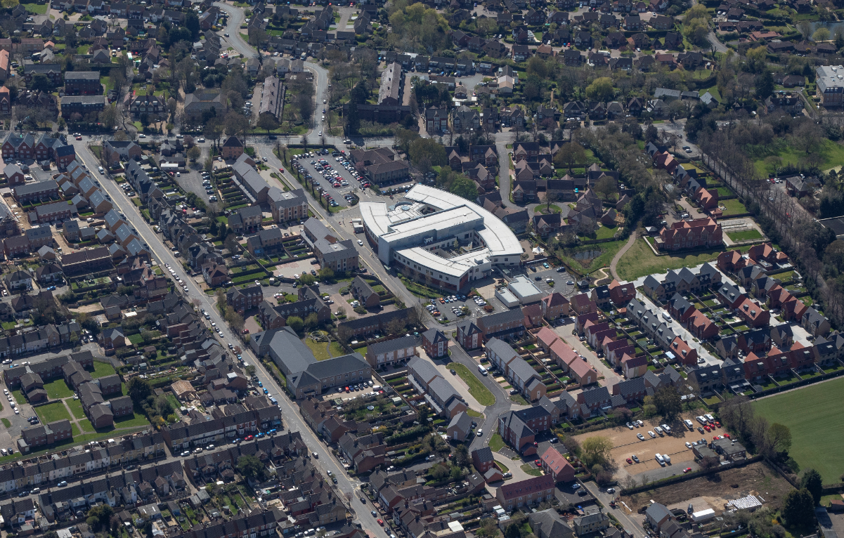Aerial view of suburban town centre with large white-roofed shopping complex, residential streets, and green spaces scattered throughout.
