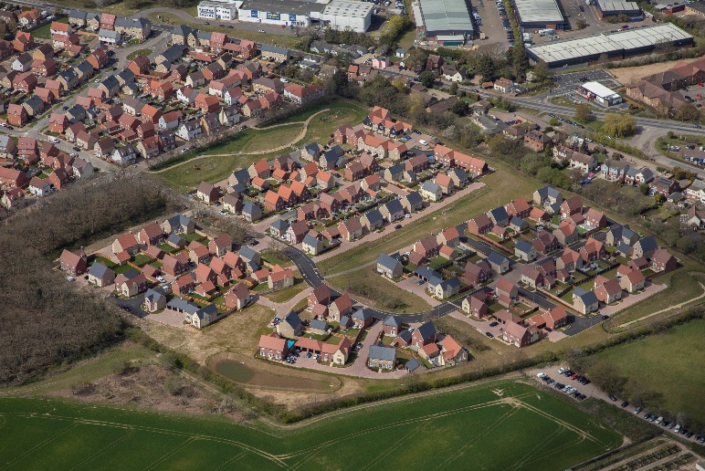 Aerial view of suburban housing estate with red-tiled roofs, curved roads, and surrounding green fields and industrial buildings.