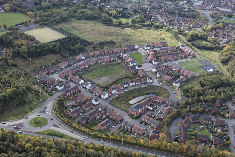 Aerial view of suburban housing estate with red-roofed houses, green fields, roads, and wooded areas surrounding residential development.