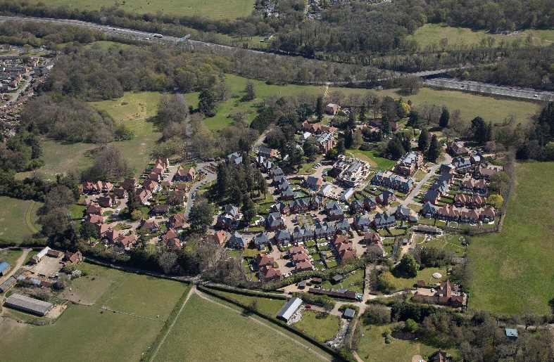 Aerial view of suburban village with houses clustered amongst trees, surrounded by green fields and roads curving through countryside.