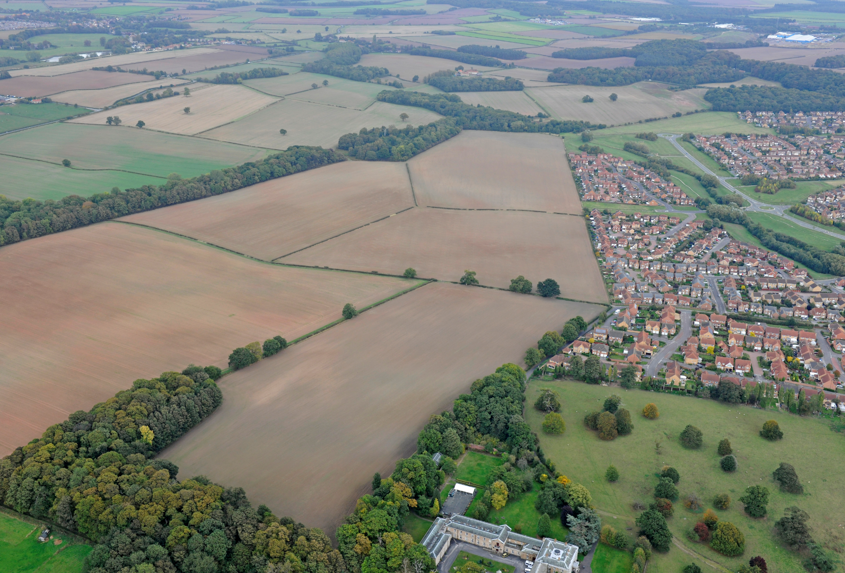 Aerial view of patchwork agricultural fields in brown and green tones with residential areas and tree lines dividing the landscape.