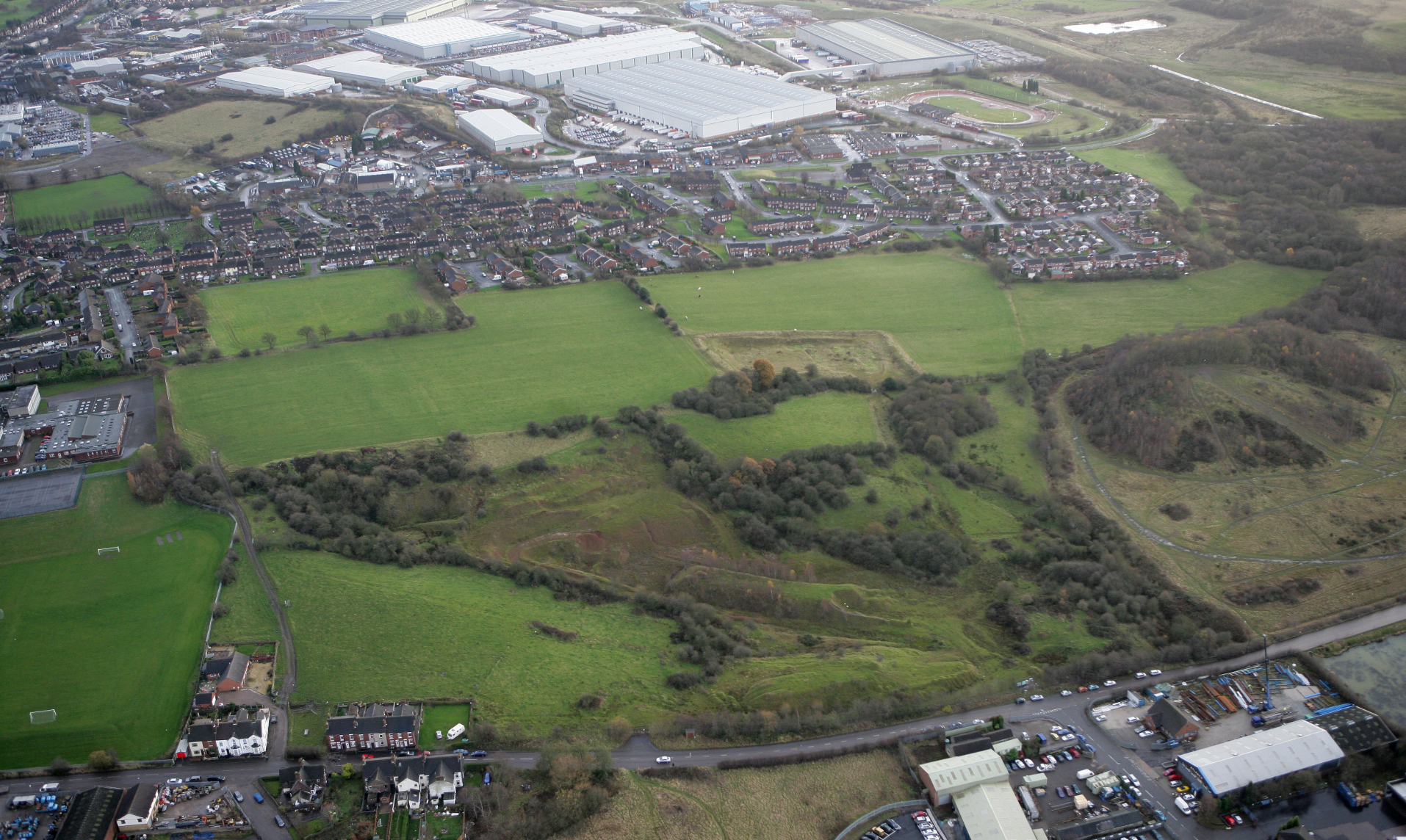 Aerial view of suburban town with green fields, residential areas, large white industrial buildings, and mixed farmland and development.