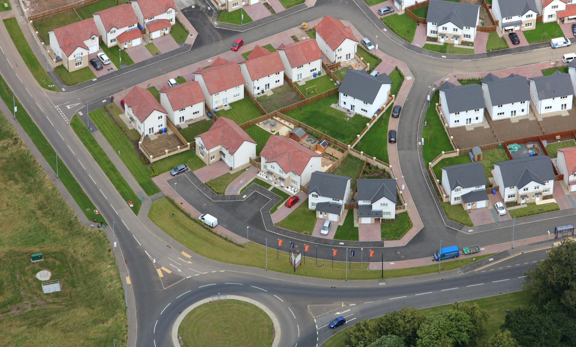 Aerial view of suburban housing estate with white and cream houses, red-tiled roofs, green lawns, and curved tarmac roads with roundabout.