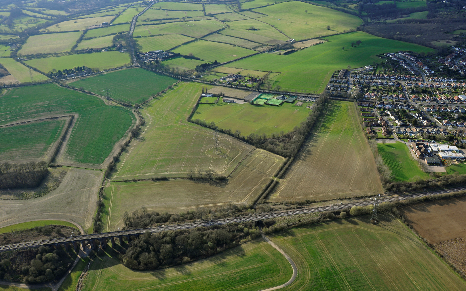 Aerial view of green agricultural fields divided by hedgerows and roads, with residential area visible in upper right corner.