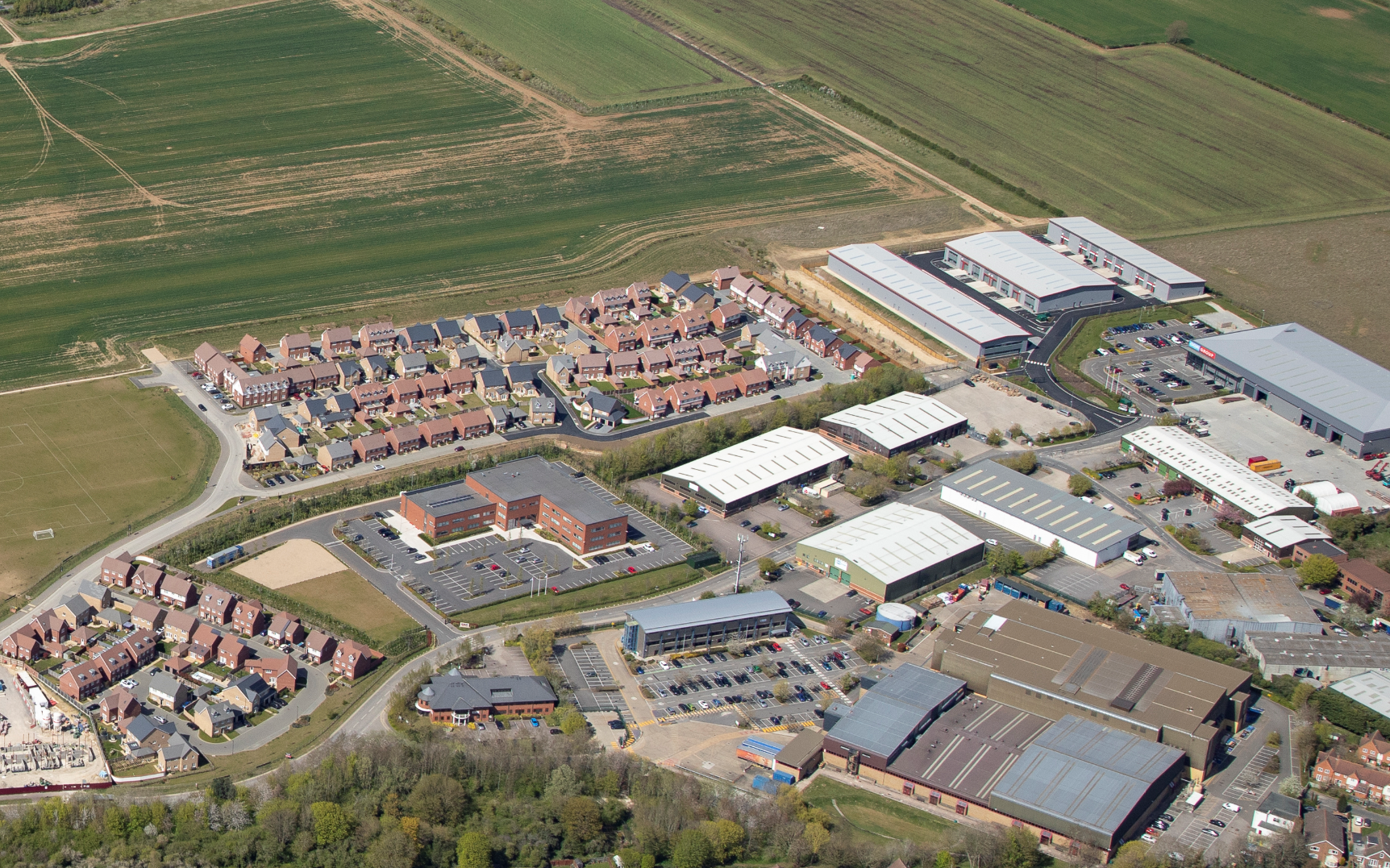 Aerial view of residential housing estate with red-roofed homes adjacent to large grey industrial buildings and warehouses surrounded by green agricultural fields.
