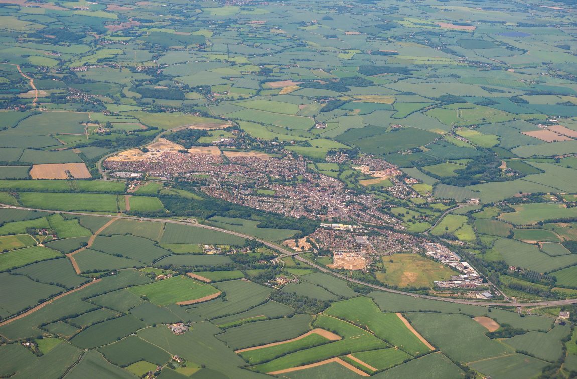Aerial view of town with red-roofed buildings surrounded by green and brown patchwork fields divided by hedgerows and roads.