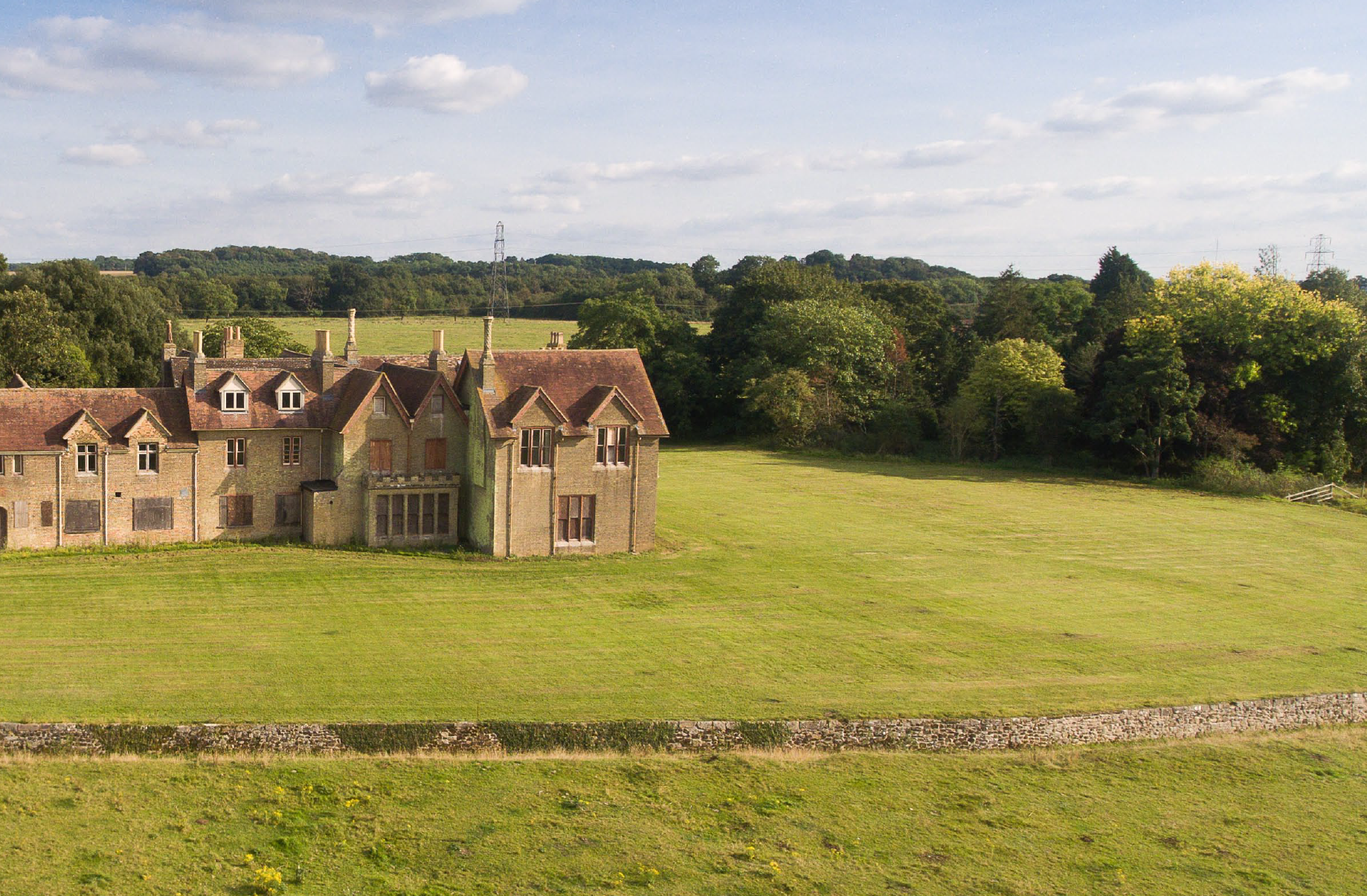 Stone manor house with mullioned windows and chimneys surrounded by green lawns, with trees and rolling hills in background.