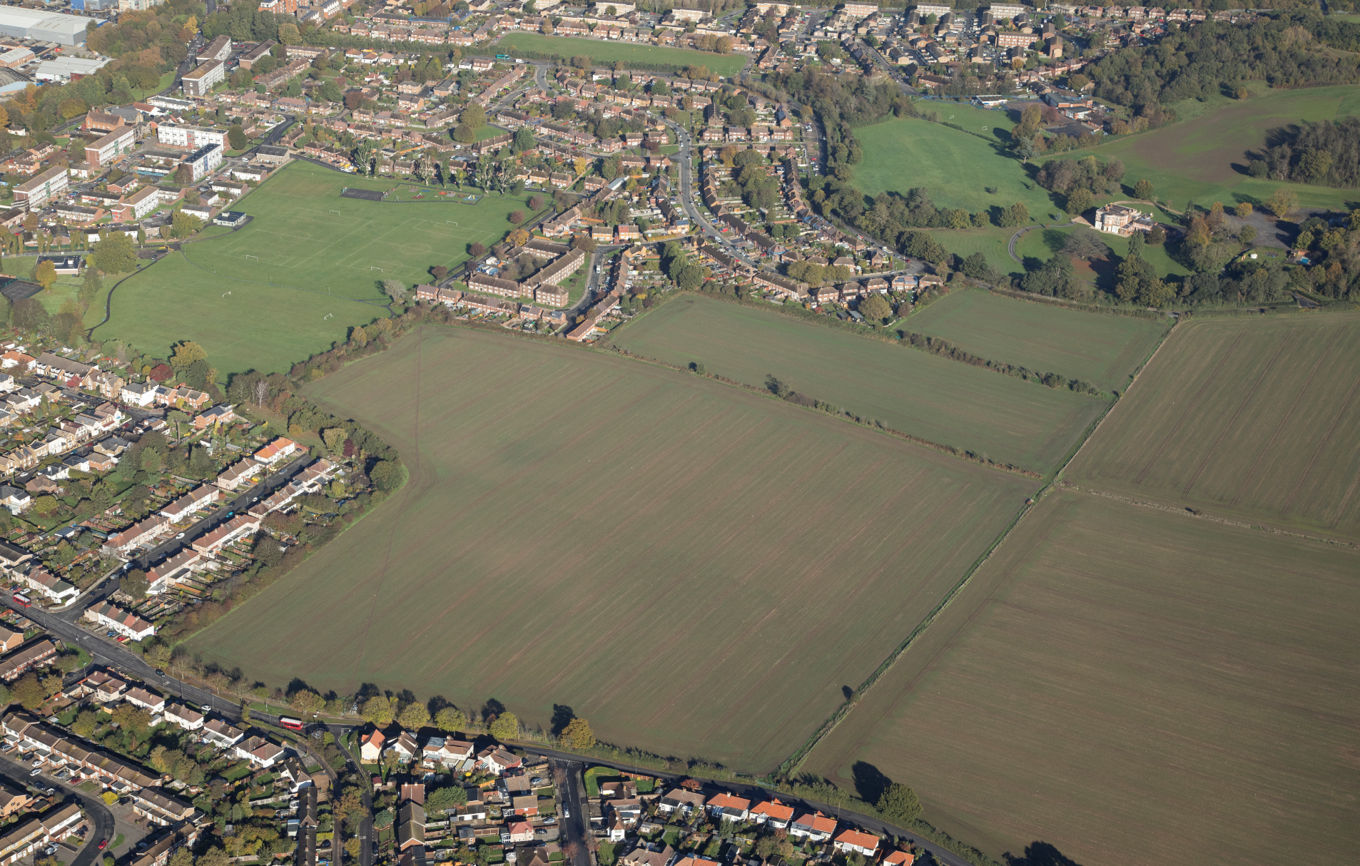 Aerial view of suburban houses surrounding large green fields with scattered trees, showing mix of residential areas and open grassland.