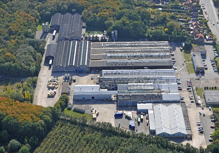 Aerial view of large industrial complex with multiple grey rectangular buildings, car parks, and surrounding green forest.
