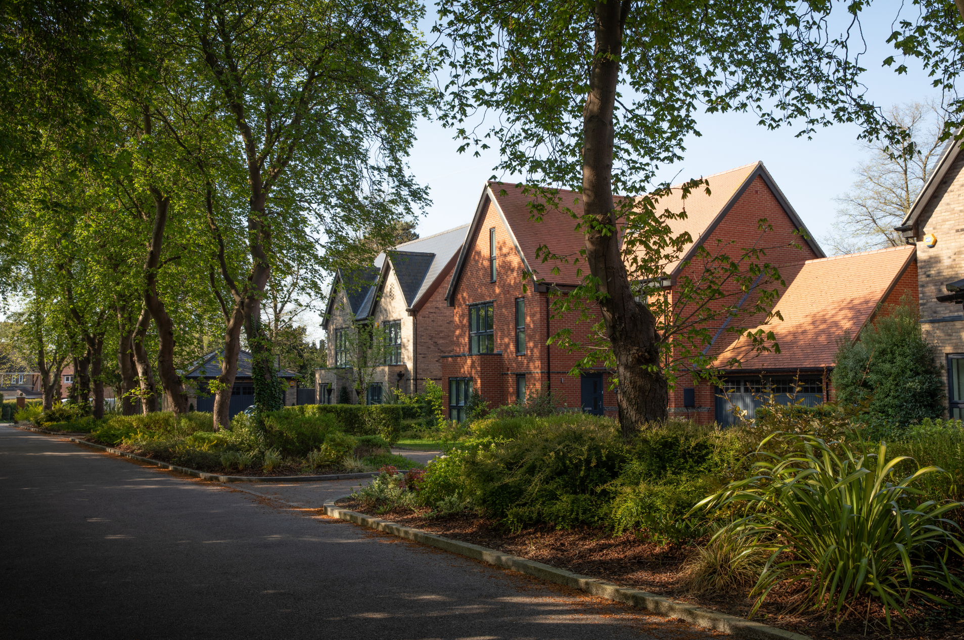 Tree-lined residential street with brick and Tudor-style houses, landscaped gardens, and overcast sky in background.