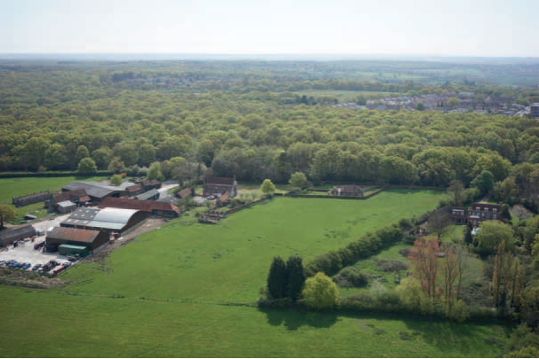 Aerial view of farm buildings with red roofs surrounded by green fields and dense woodland stretching to horizon under cloudy sky.
