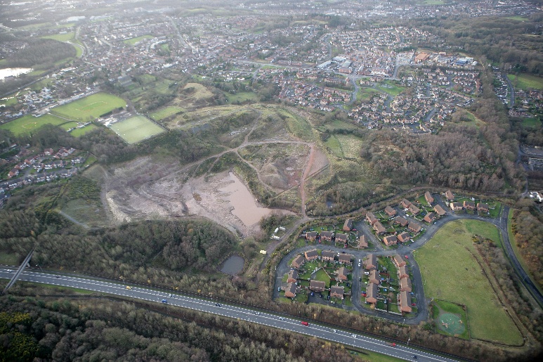 Aerial view of suburban area with motorway, residential estates, green fields, and large quarry or construction site with exposed earth.